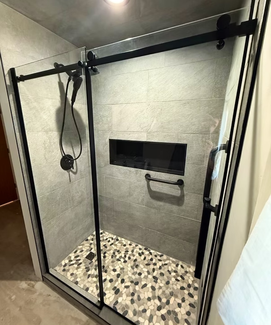 Walk-in shower with gray tile walls, a pebble tile floor and a black-framed glass door, built by DRUX Tech